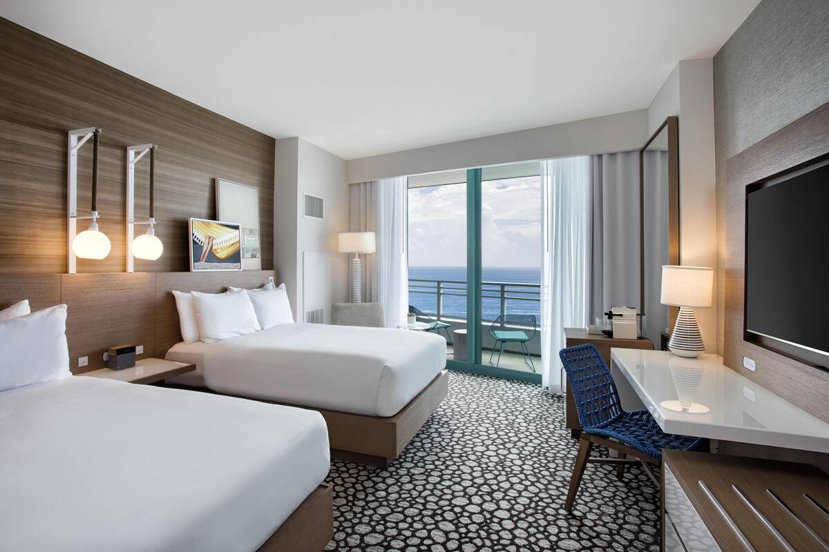 A hotel room with two beds, a desk, a chair, a TV, and a balcony with a view of the ocean.
