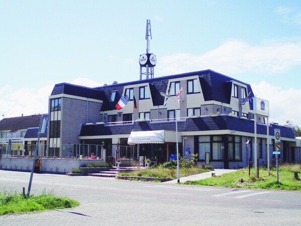 Un hotel con diseño arquitectónico moderno, que destaca una torre del reloj prominente, varias banderas y un cielo despejado.