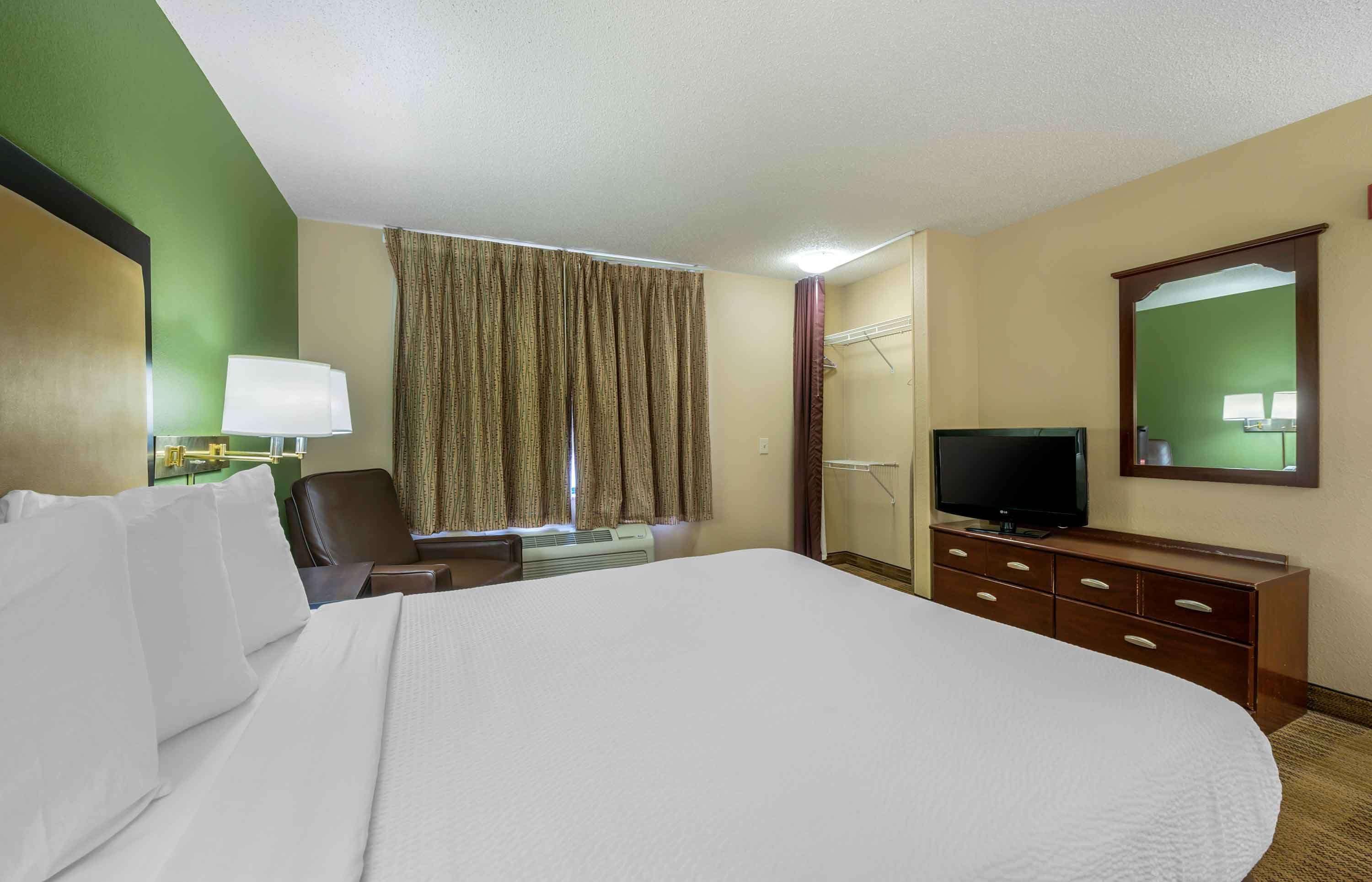 A hotel room with a large bed, a television, a desk with a lamp, and a window with curtains.