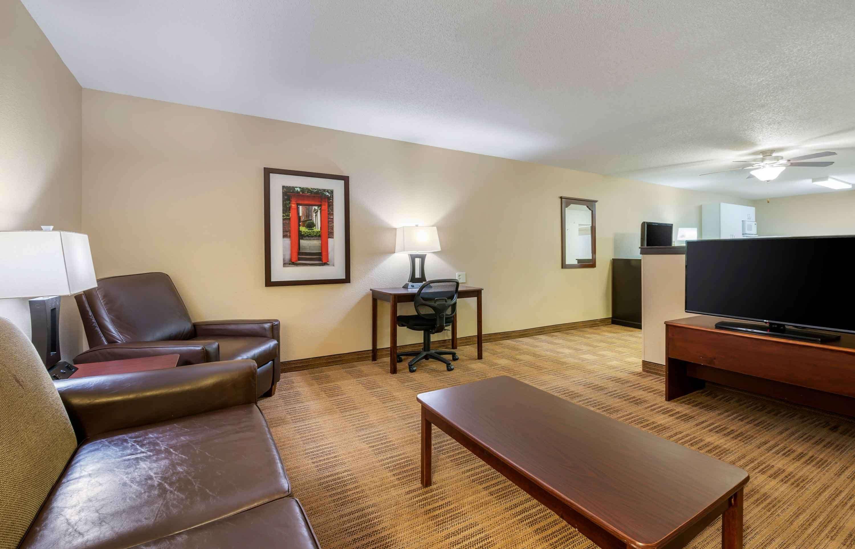 A hotel room with a leather sofa, a wooden coffee table, a desk with a chair, a television, and a lamp.