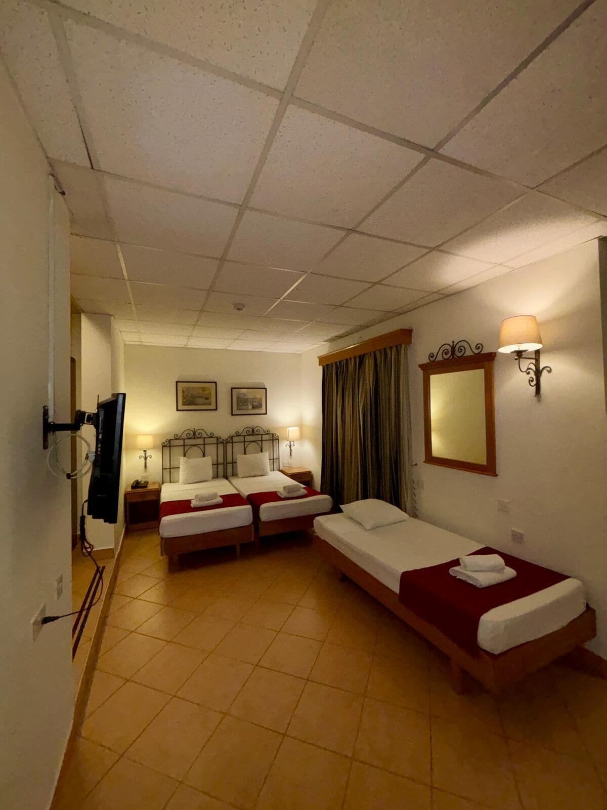 Habitación de hotel con dos camas individuales, una cama más grande, un televisor, un espejo y una lámpara.