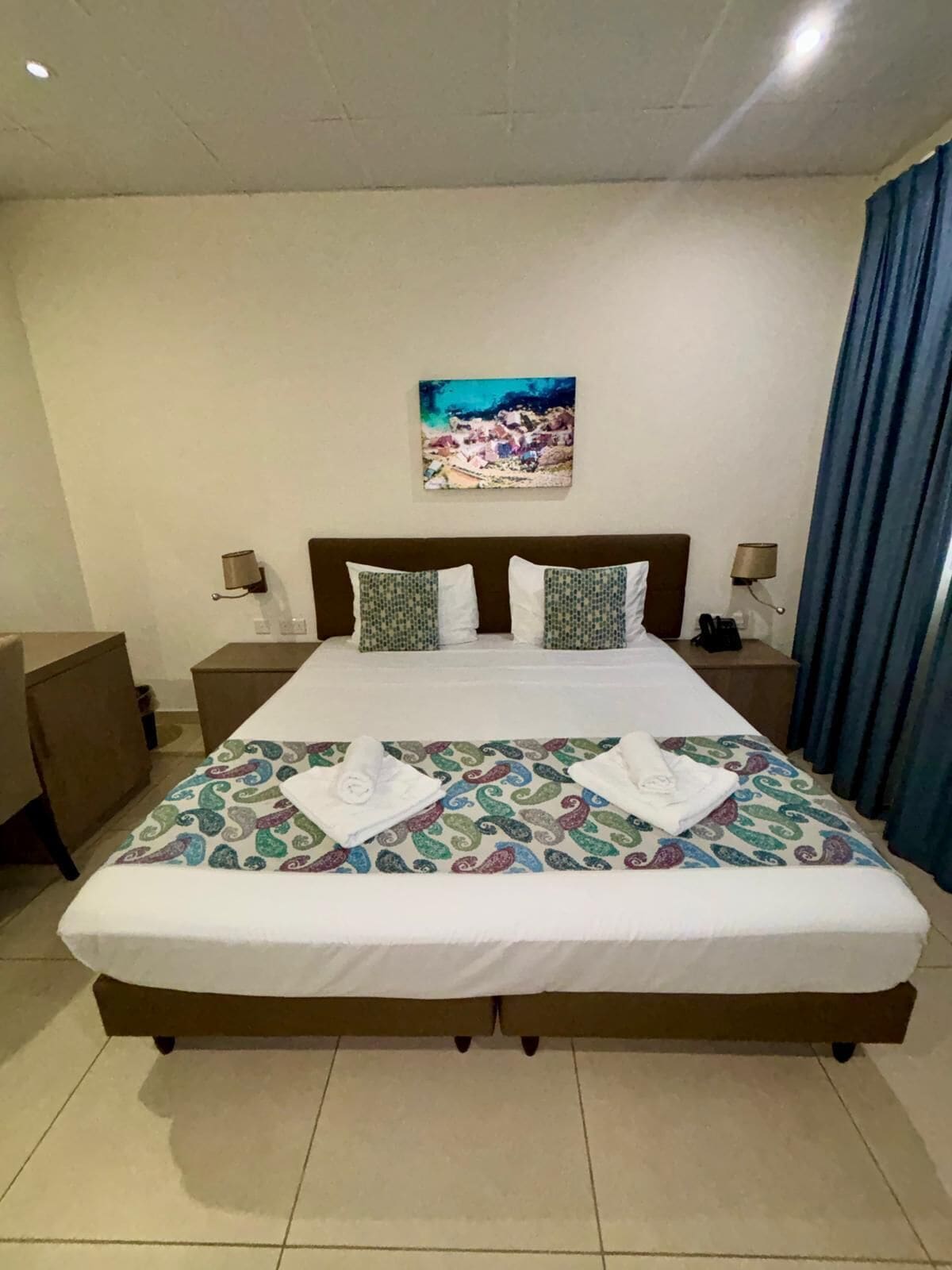 Une chambre d’hôtel avec un grand lit, deux oreillers, une table de chevet, une lampe, un tableau au mur et un rideau bleu.