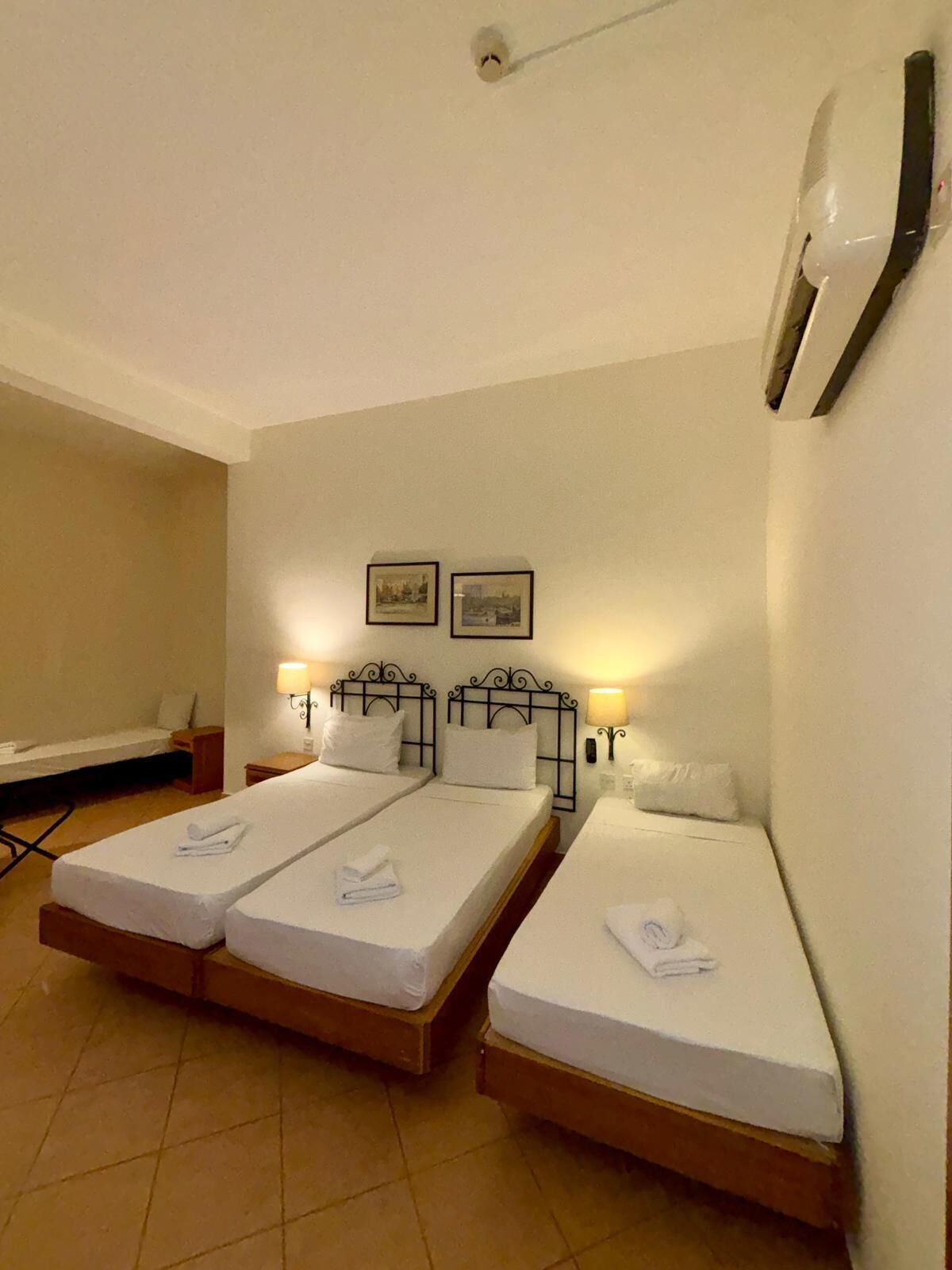 A hotel room with two single beds, a small bedside table with a lamp, a wall-mounted air conditioner, and a framed picture above the beds.