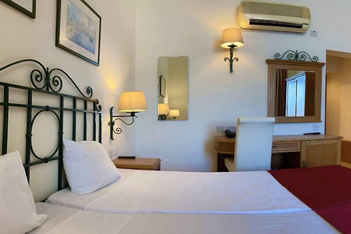 A hotel room with a bed, a desk, a chair, a wardrobe, a mirror, and an air conditioning unit.
