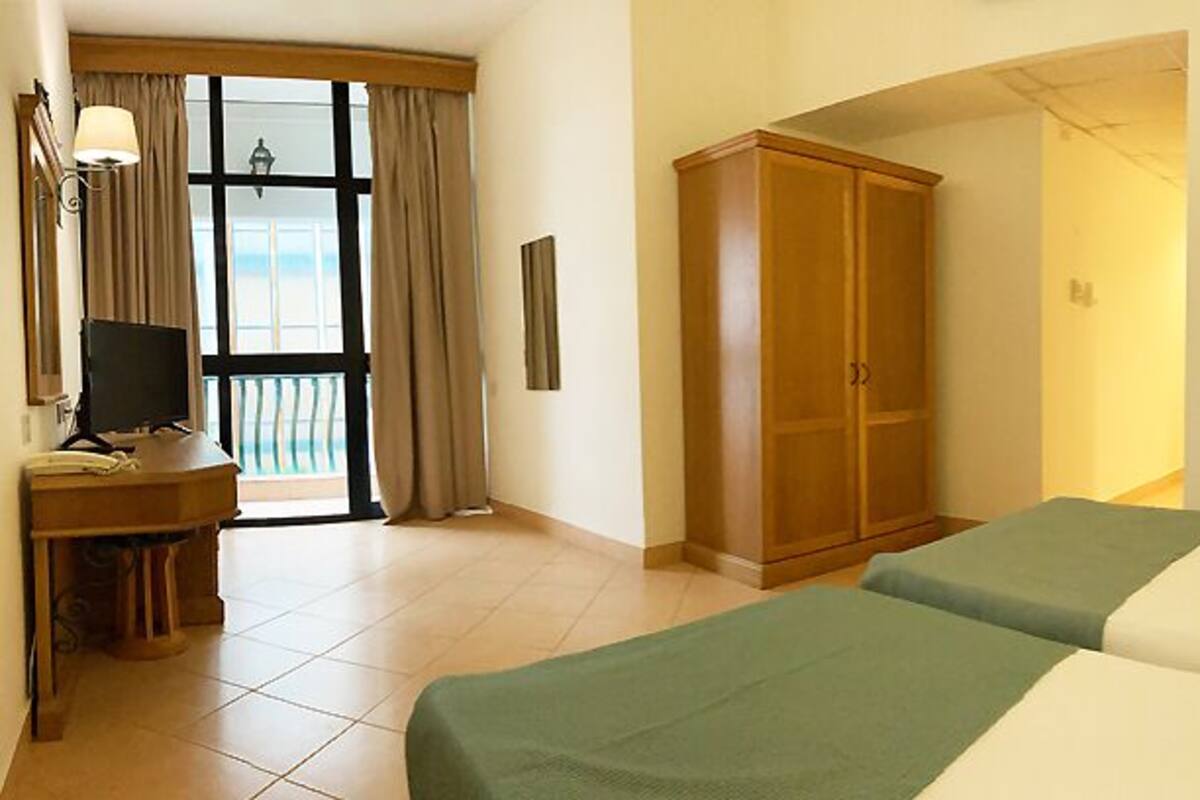 A hotel room with two beds, a desk with a TV, a wardrobe, and a balcony with curtains.