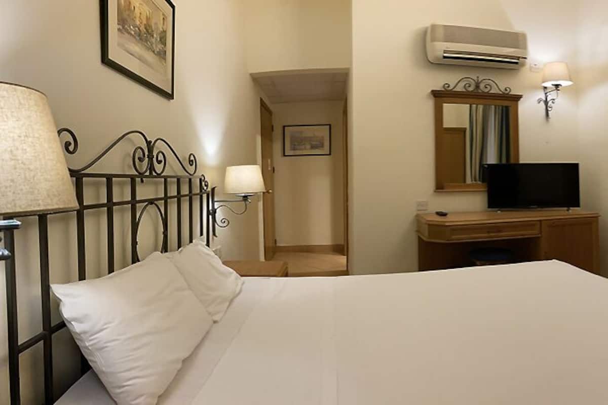A hotel room with a bed, a desk with a television, a lamp, an air conditioner, and a window with curtains.