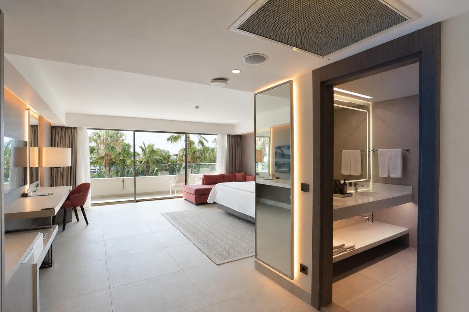 A modern hotel room with a bed, a desk, a chair, a large window with a view, and a bathroom with a mirror and towel rack.