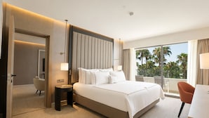 A hotel room with a large bed, a desk, a chair, and a balcony with a view of palm trees. -  Mirage Park Resort - Premium All Inclusive (Kemer)