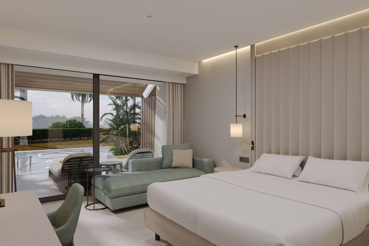 A modern hotel room with a large bed, a seating area with a chair and a small table, and a view of a pool and palm trees through a glass door.