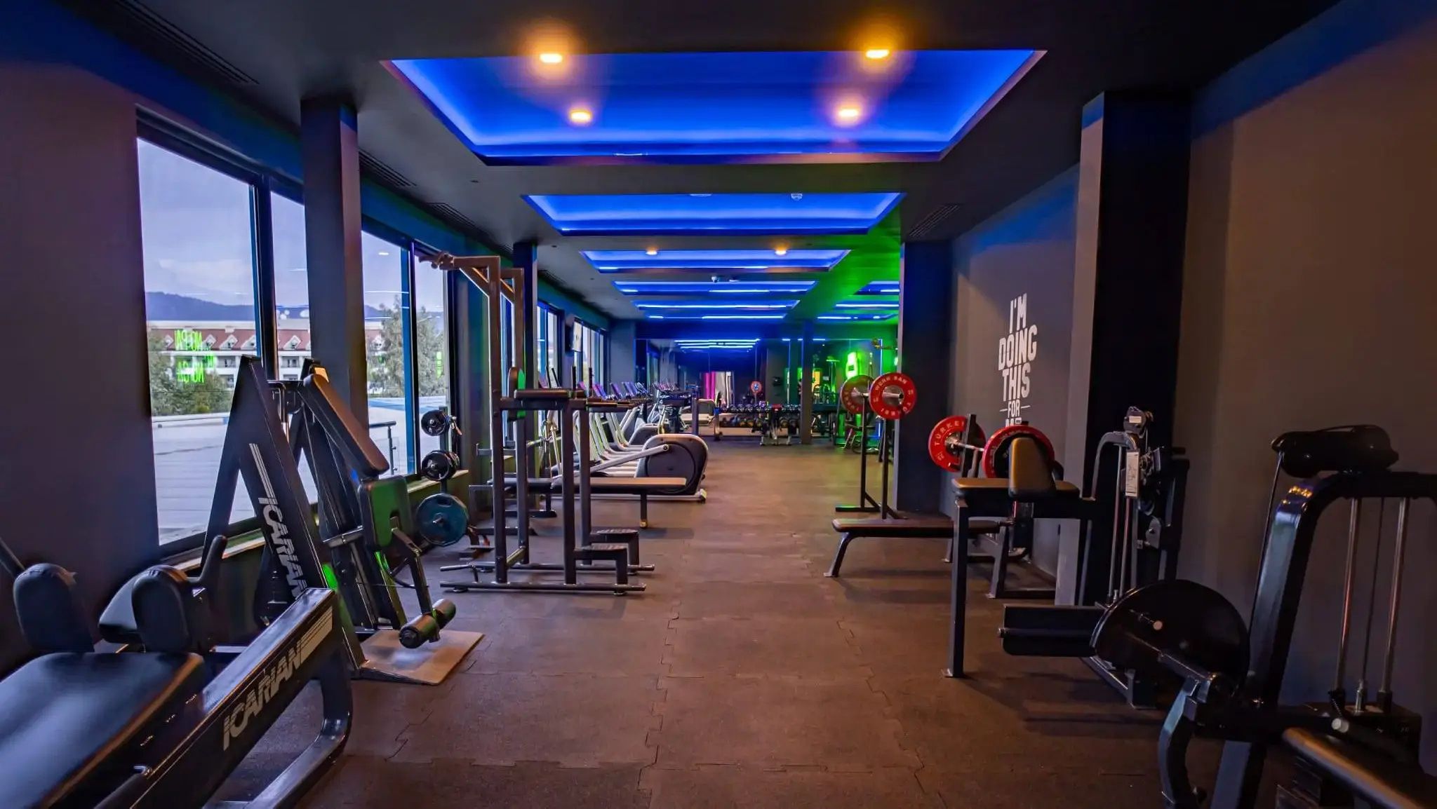 Una palestra con diverse attrezzature per esercizi, ampie finestre e una citazione motivazionale sulla parete.