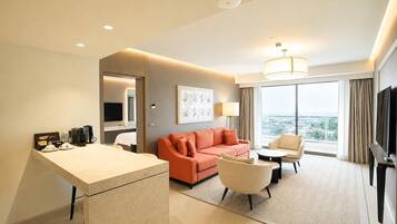 A hotel room with a sofa, armchair, TV, and a desk with coffee maker and cups.