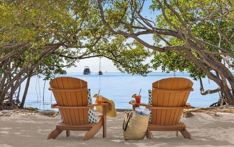 Two wooden Adirondack chairs on a sandy beach with a view of the sea and a sailboat in the distance.
