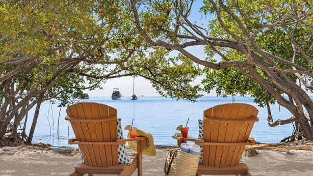 Dos sillas Adirondack de madera sobre una playa arenosa, con vista al mar y un velero a lo lejos.