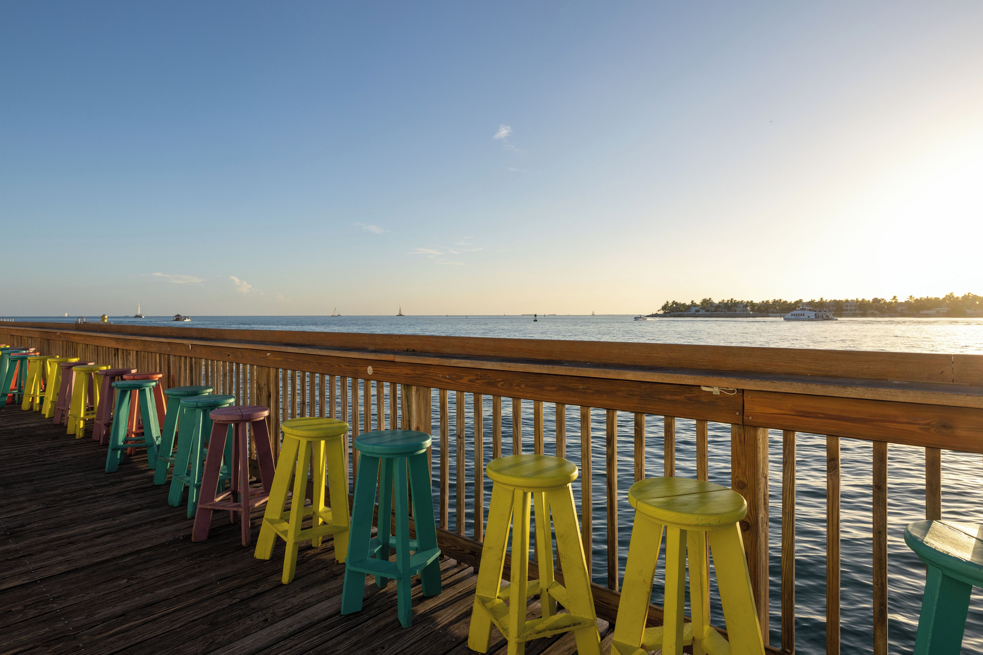 A wooden deck with colorful stools and a wooden railing overlooking a calm body of water.