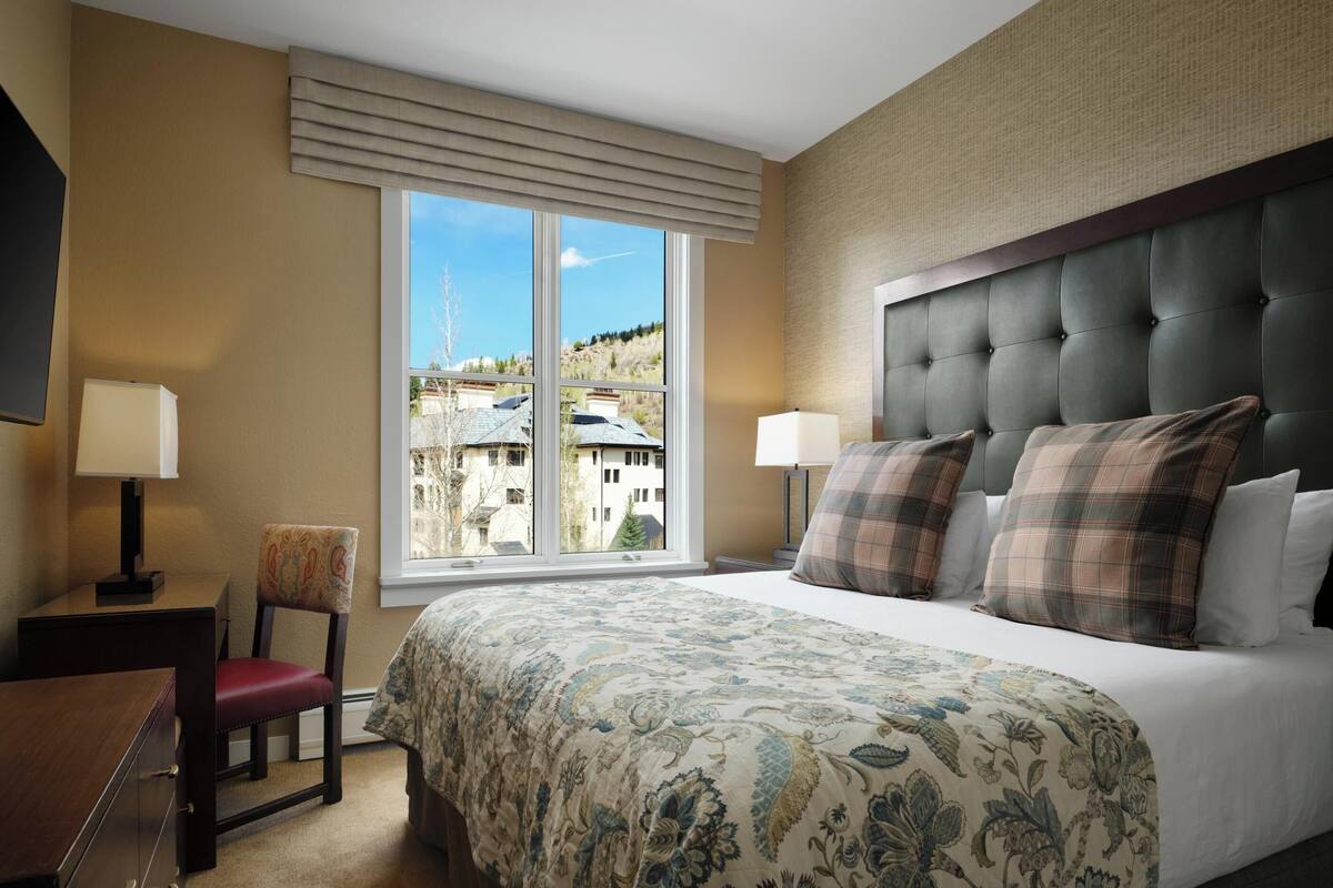 A hotel room with a large bed, two bedside lamps, a chair, a desk, and a window with a view of buildings.