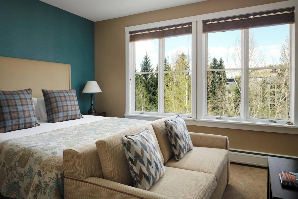 A bedroom with a bed, a sofa, a window with a view of trees, and a lamp.