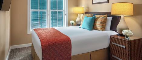 A hotel room with a large bed, two bedside lamps, a nightstand, and a window with blinds.