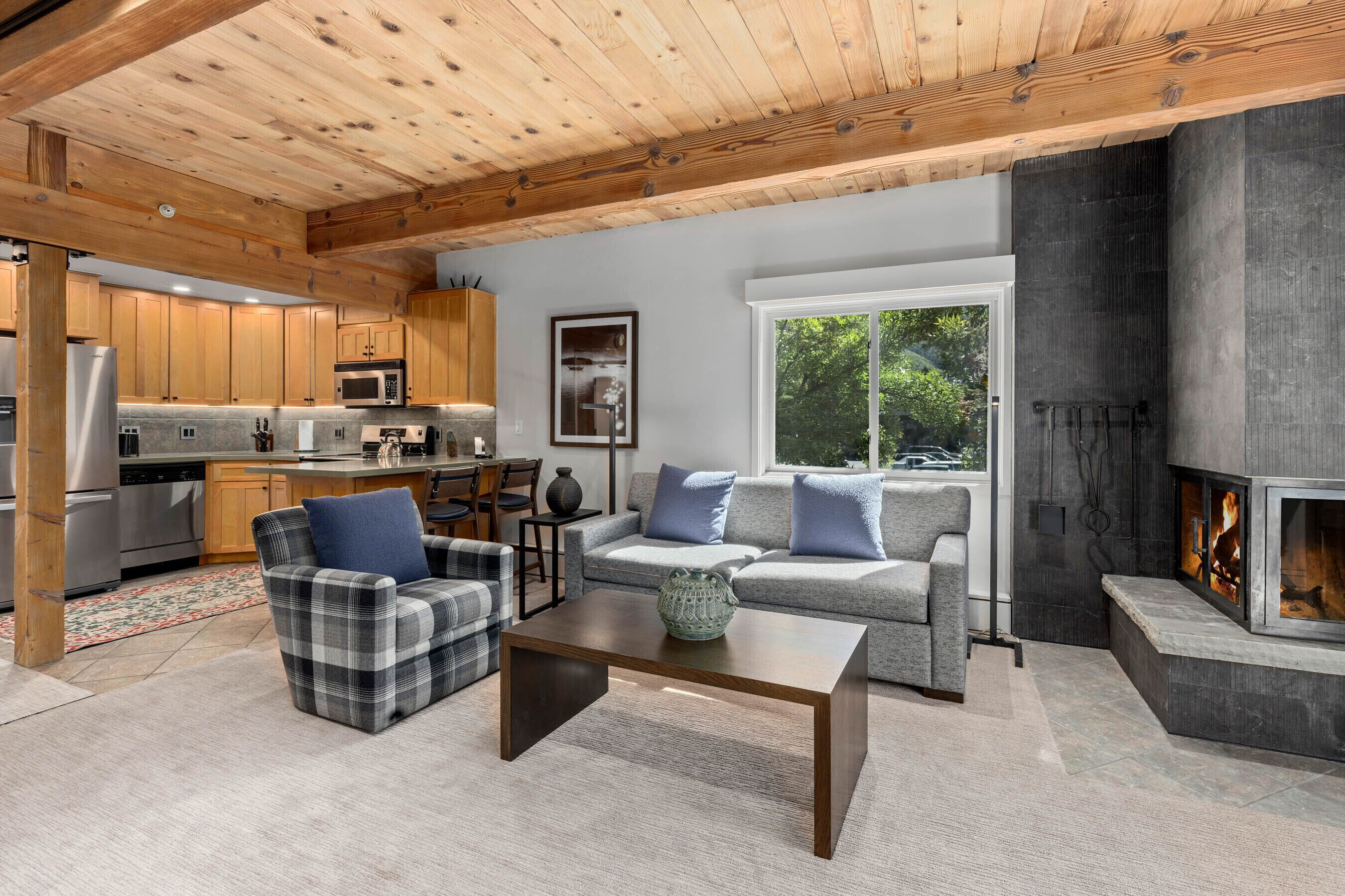A modern living room with a fireplace, wooden ceiling, and a kitchen in the background.