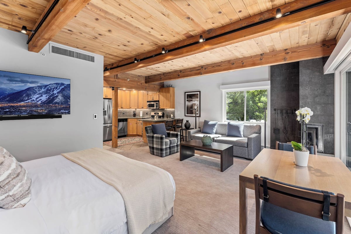 A spacious living area with a wooden ceiling, a large flat-screen TV, a sofa, a dining table, and a kitchen in the background.