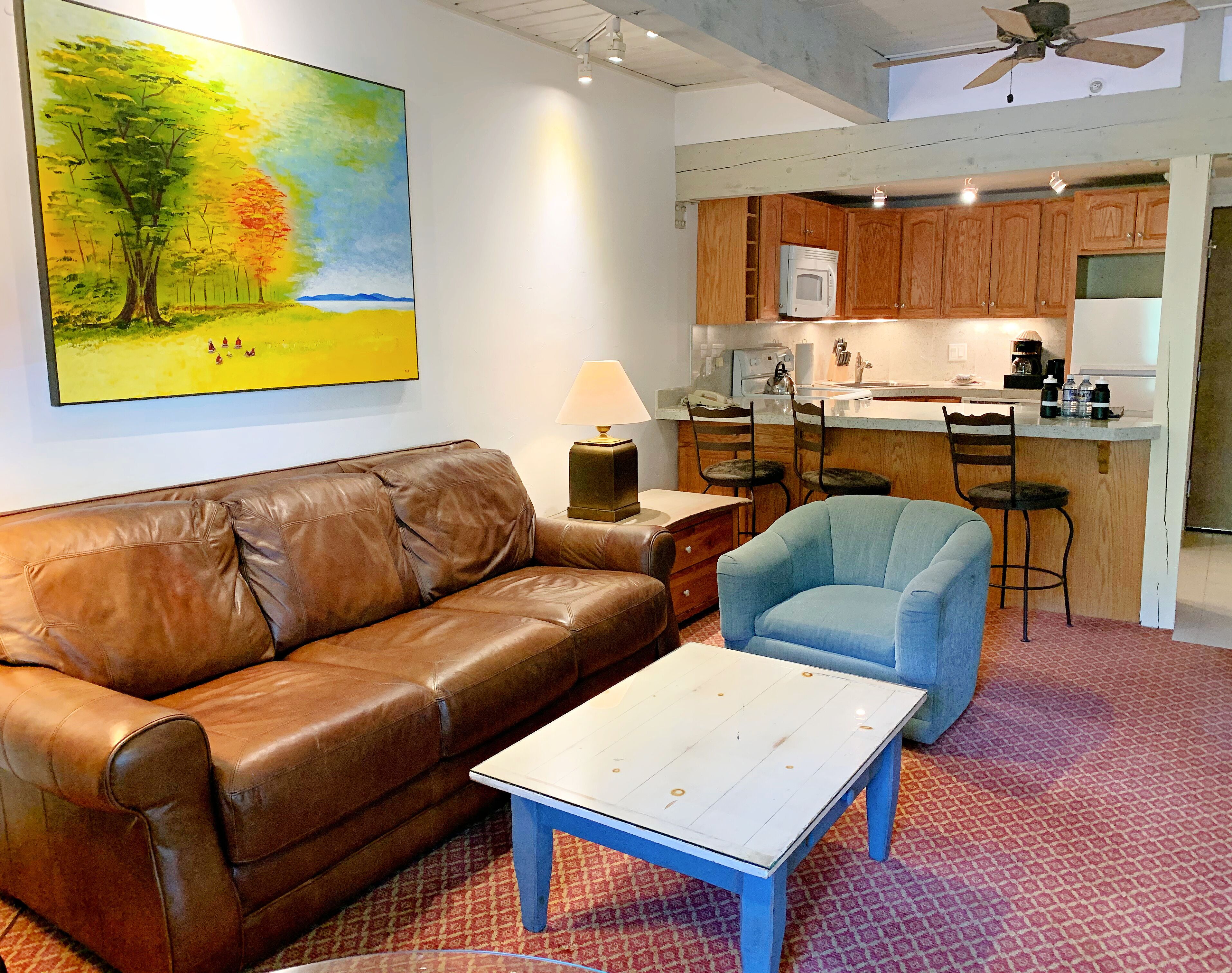 A living room with a brown leather couch, a blue armchair, a white coffee table, a painting of a landscape, and a kitchen area with wooden cabinets and a microwave.