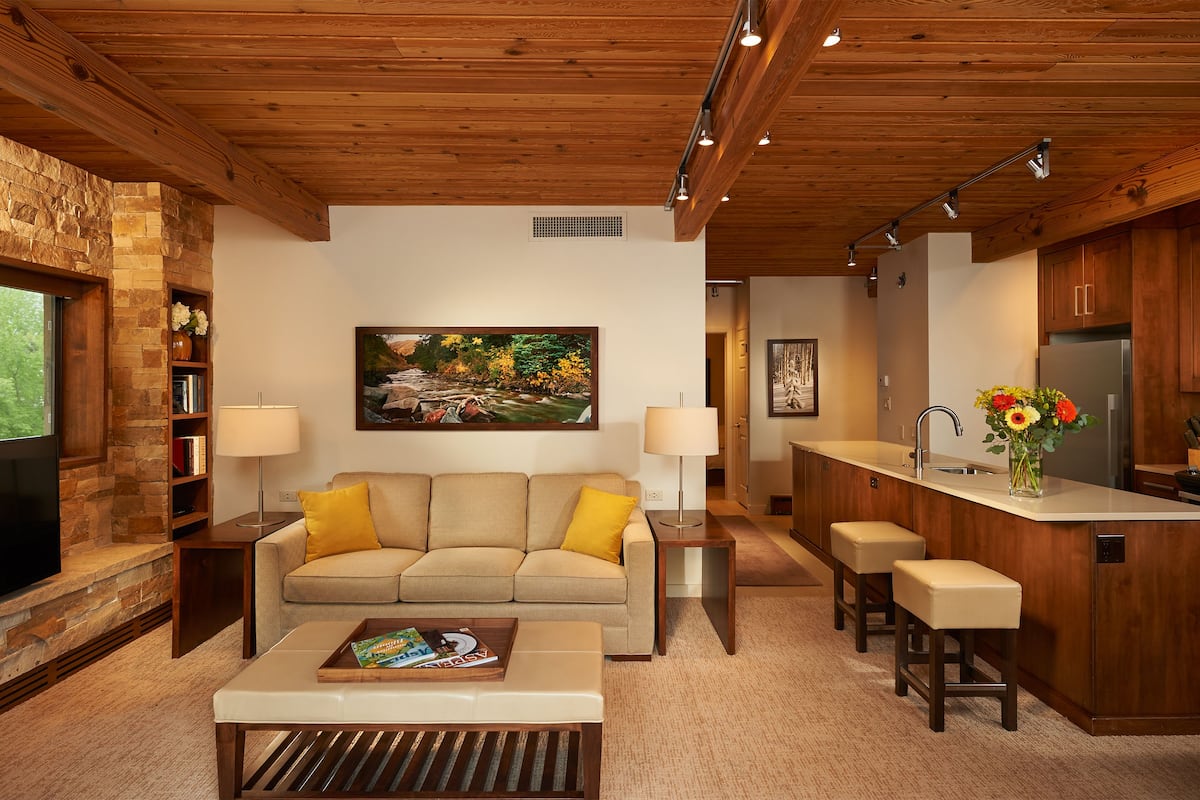 A modern living room with a sofa, coffee table, and a painting on the wall. There is a kitchen area with a bar and stools.