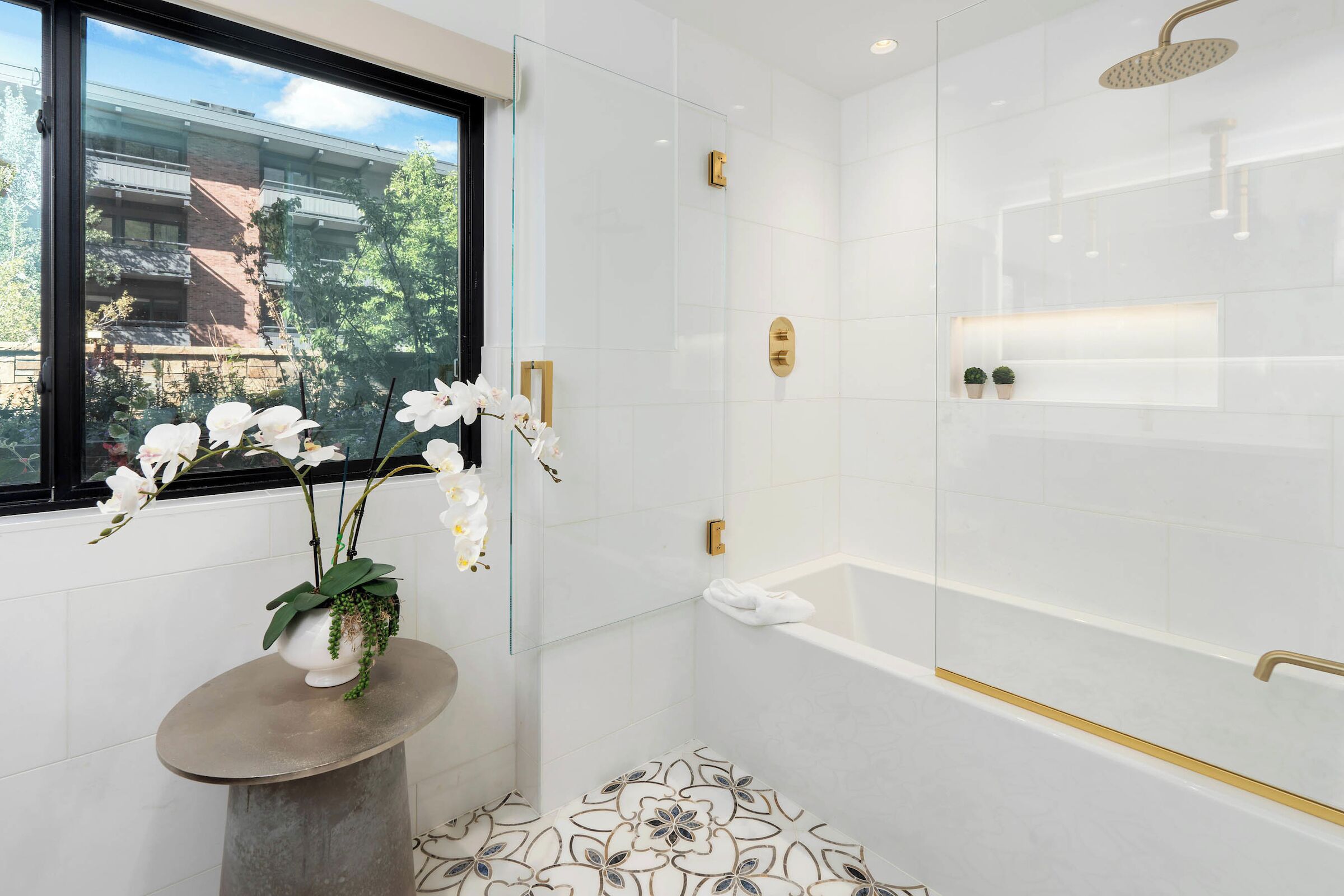 A modern bathroom with a glass shower enclosure, a bathtub with gold fixtures, and a window with a view of buildings and trees.