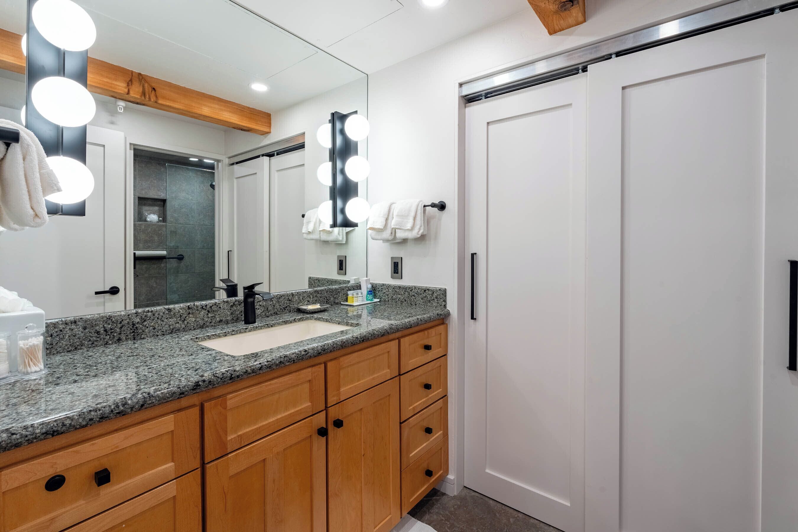 A modern bathroom with a granite countertop, wooden cabinets, a large mirror with lights, and a white door.