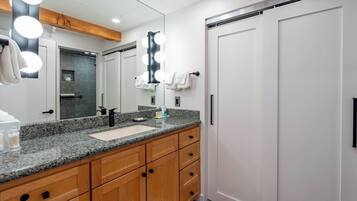 A modern bathroom with a granite countertop, wooden cabinets, a large mirror with lights, and a white door.