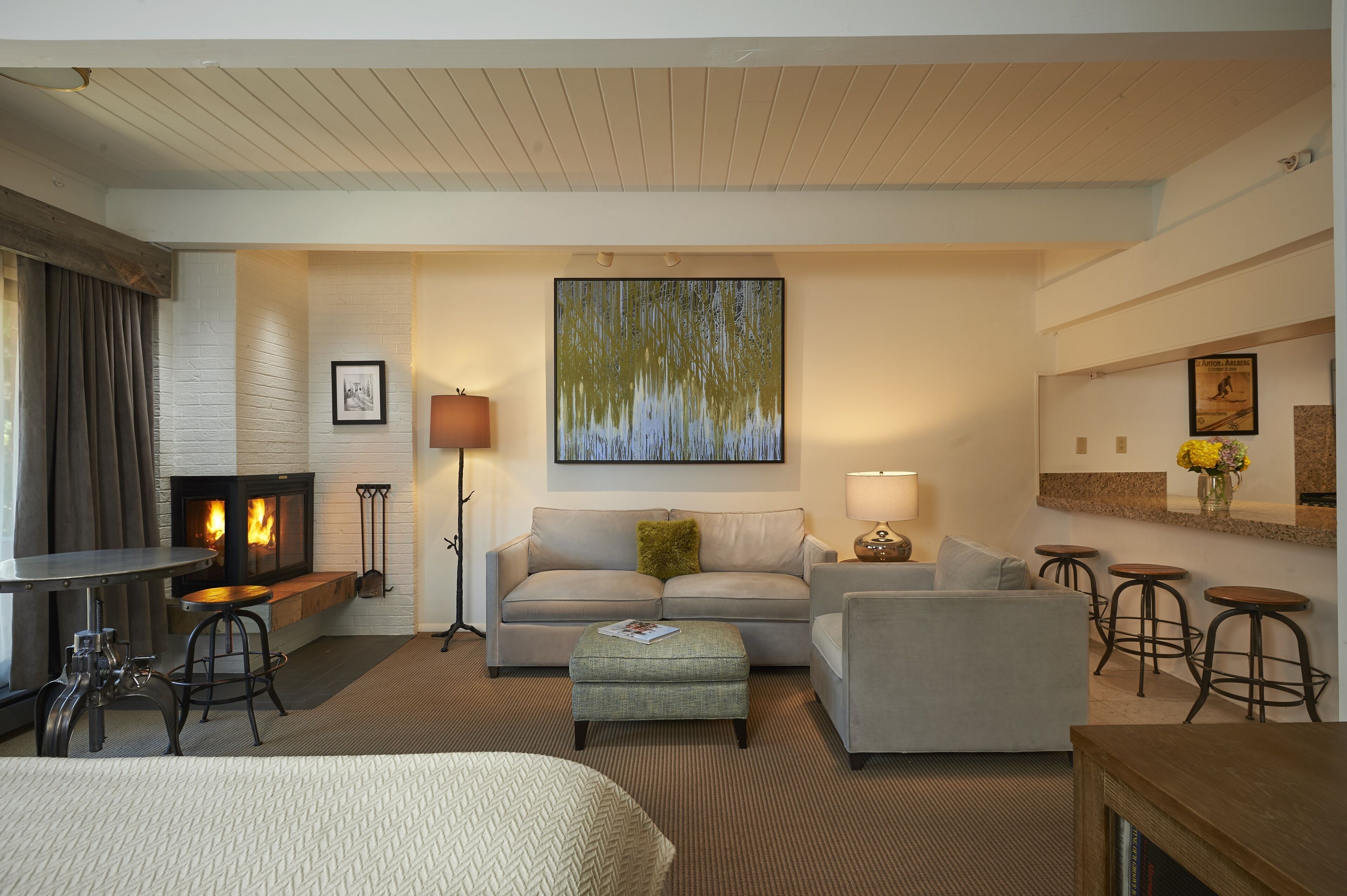 A hotel room with a fireplace, a sofa, a coffee table, a small ottoman, a bar area with stools, and a large painting on the wall.