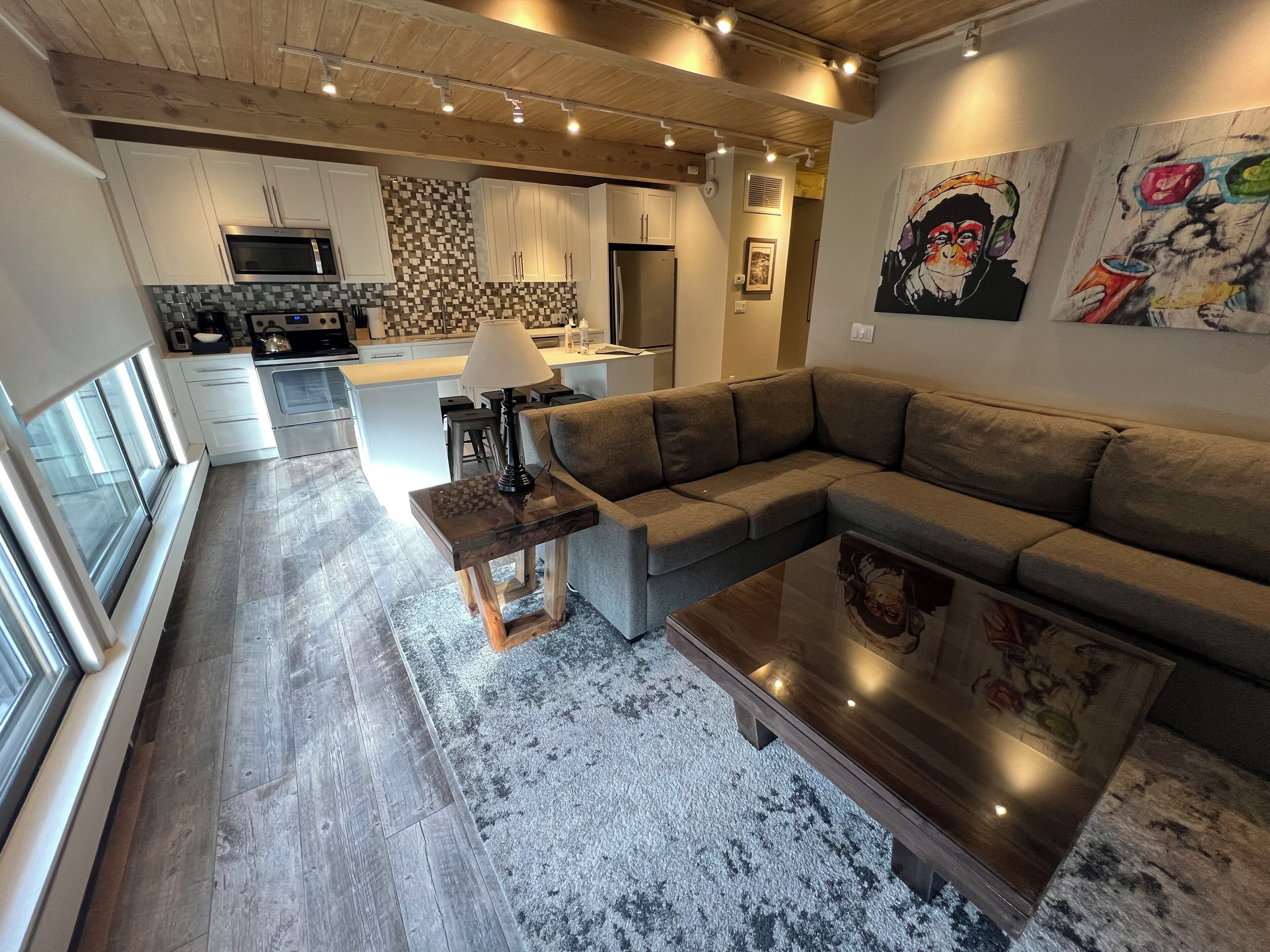 A modern kitchen with white cabinets, a microwave, and a tiled backsplash. There is a dining area with a gray sofa and a coffee table. The room has a wooden ceiling and a large window.