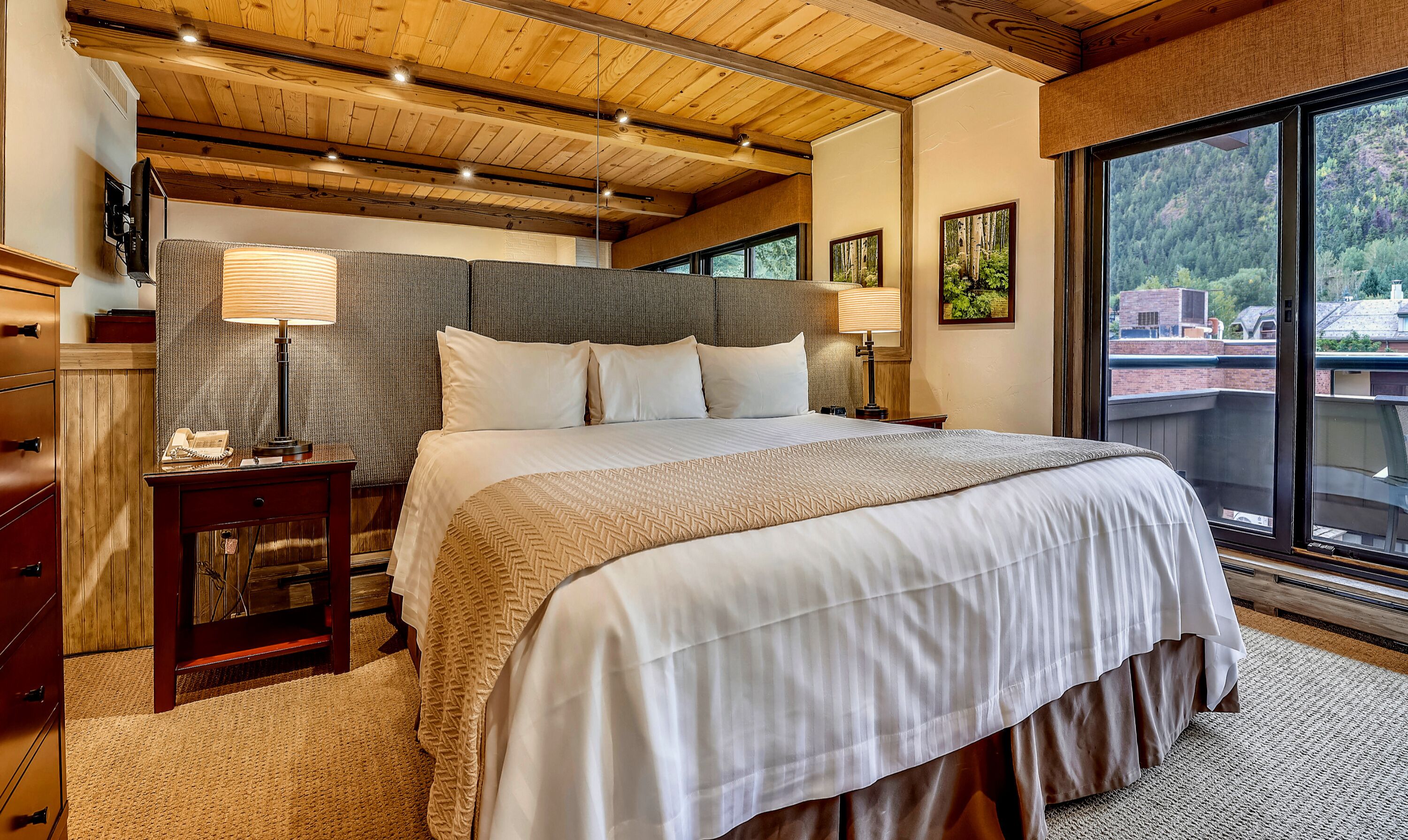 A bedroom with a large bed, wooden ceiling beams, a nightstand with a lamp, and a view of a forest through a large window.