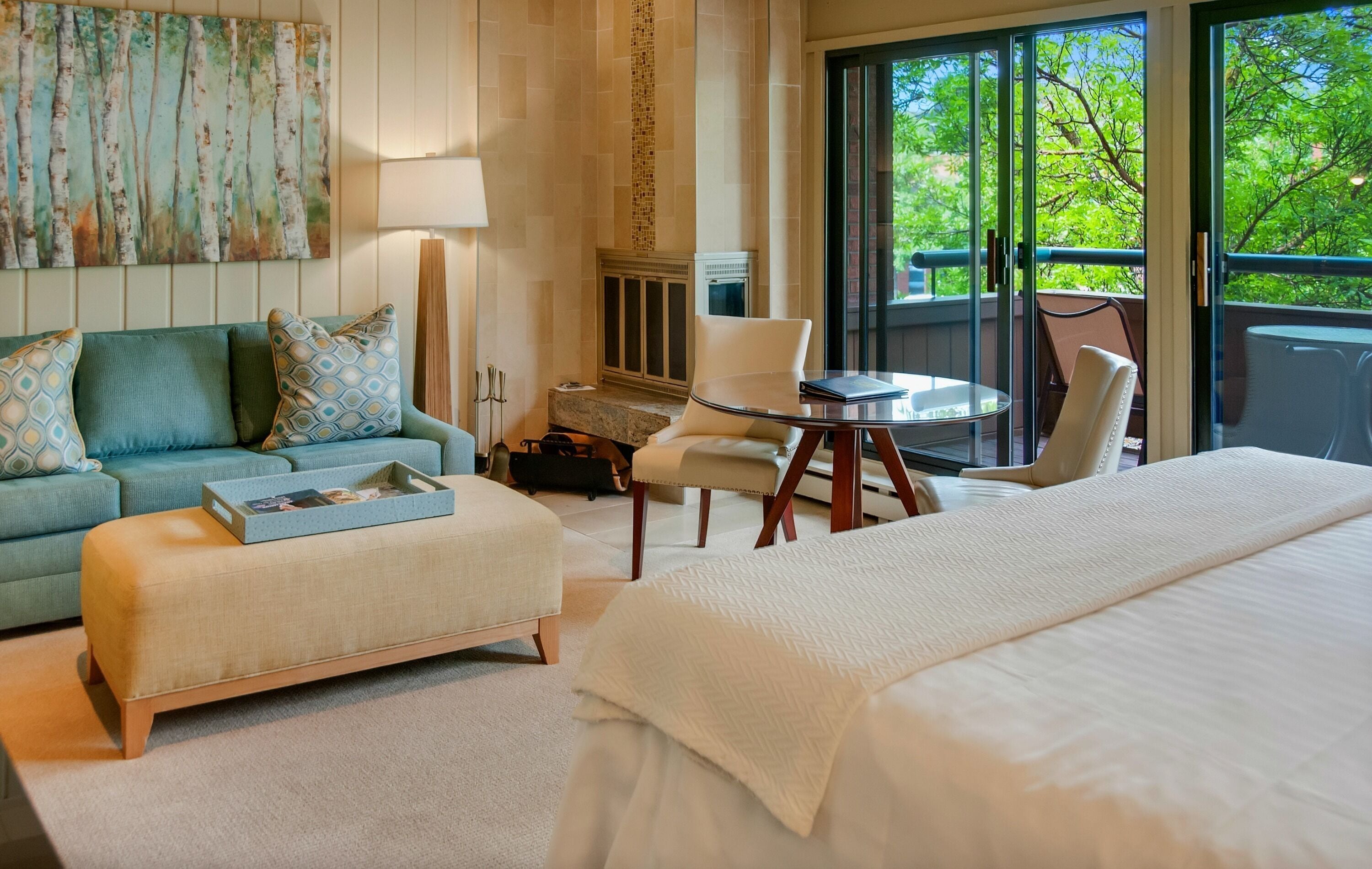 Une chambre d’hôtel avec un grand lit, un canapé, une petite table et une chaise. On y trouve une cheminée et, par les fenêtres, on peut admirer la vue sur les arbres.