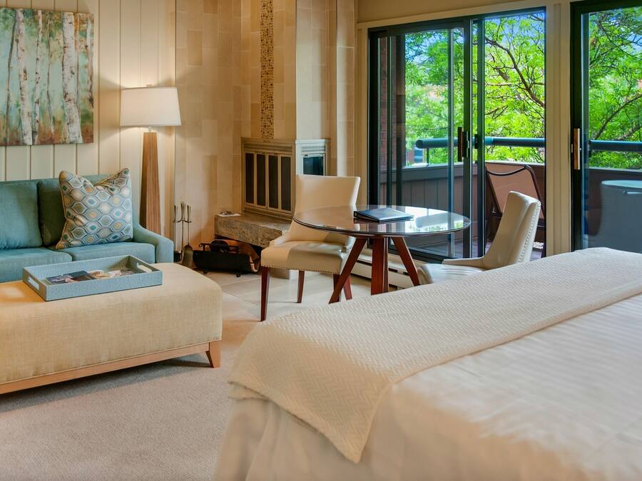 A hotel room with a large bed, a sofa, a small table, and a chair. There is a fireplace and a view of trees through the windows.