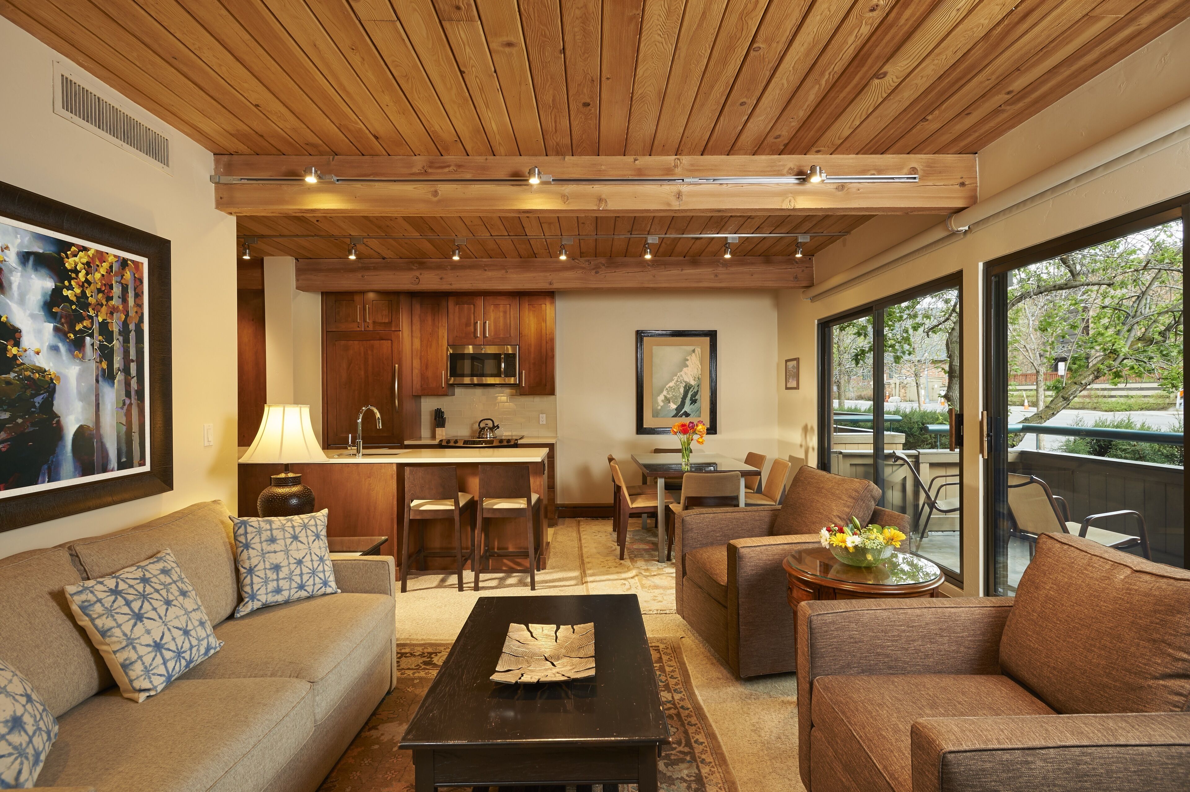 A spacious living area with a wooden ceiling, a kitchen, and large windows offering a view of the outdoors.