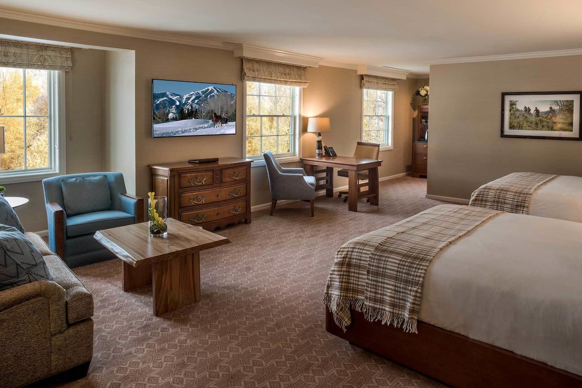 A hotel room with two beds, a desk, a chair, a TV, and a window with a view of mountains.