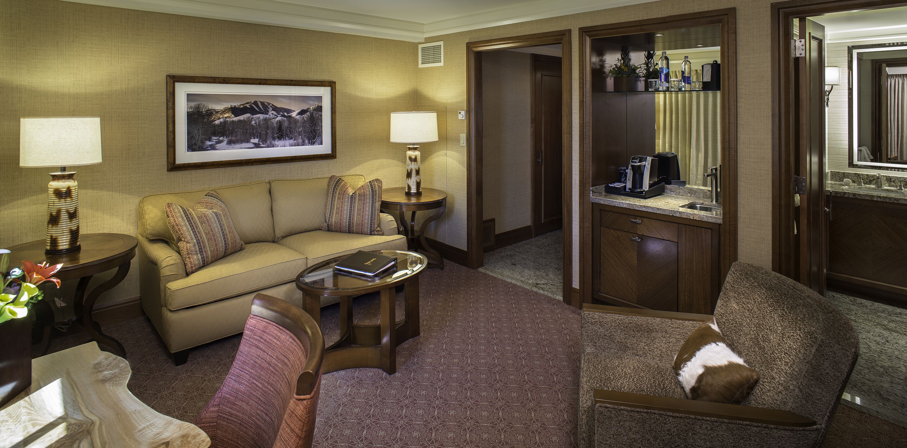 Habitación de hotel con sofá, sillón, mesa de centro, lámparas, un cuadro enmarco y minibar.