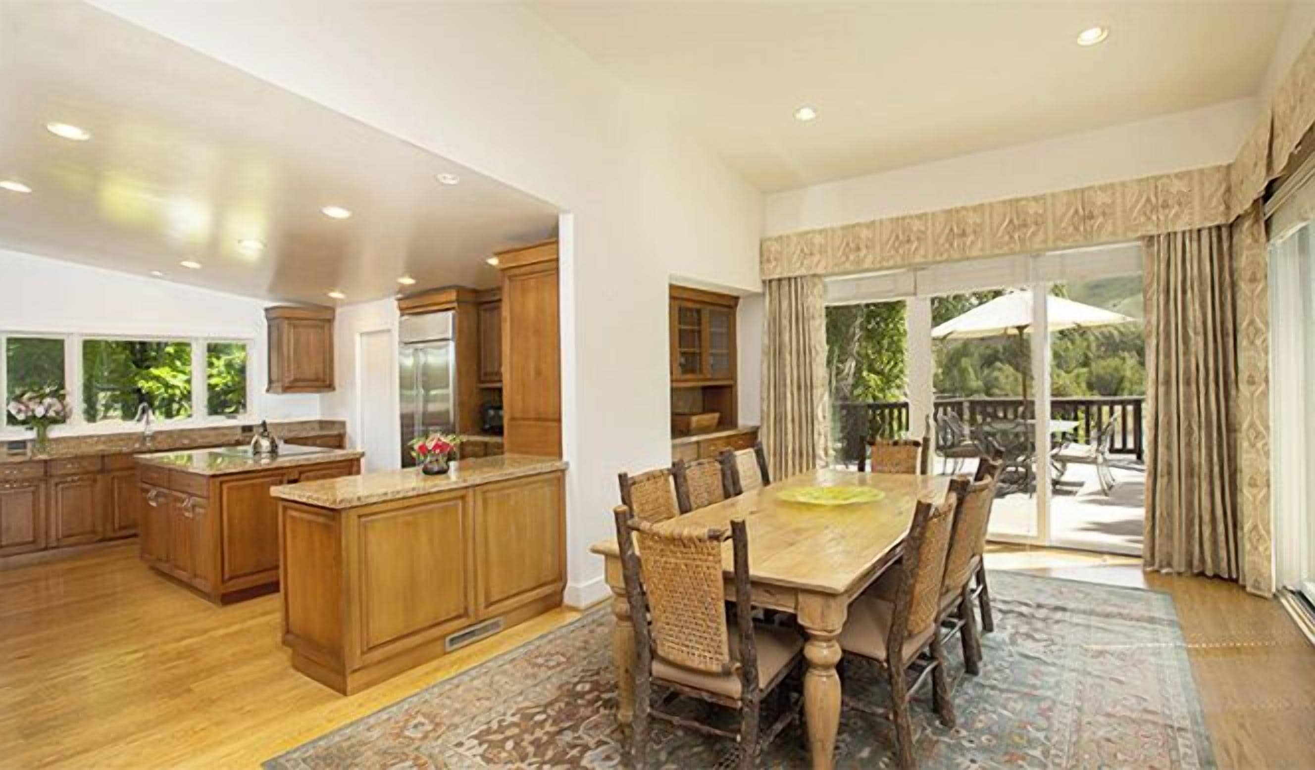 Una cocina amplia con armarios de madera y una isla grande, un comedor con mesa y sillas de madera, y vistas a un jardín exuberante a través de una puerta corrediza de vidrio.