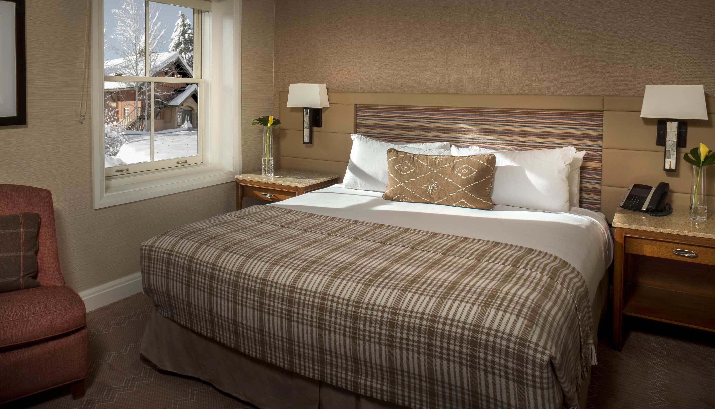 Habitación de hotel con una cama grande, dos mesitas de noche, una silla y una ventana con vistas a un paisaje nevado.