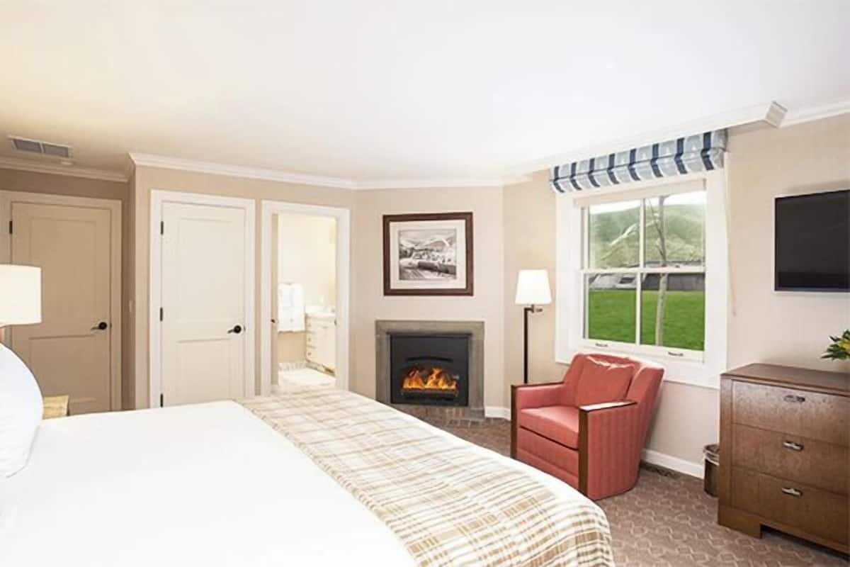A hotel room with a bed, a fireplace, a red chair, a TV, and a window with a view of the outdoors.