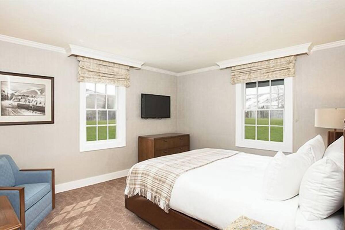 A bedroom with a bed, a chair, a dresser, a television, and two windows with curtains.