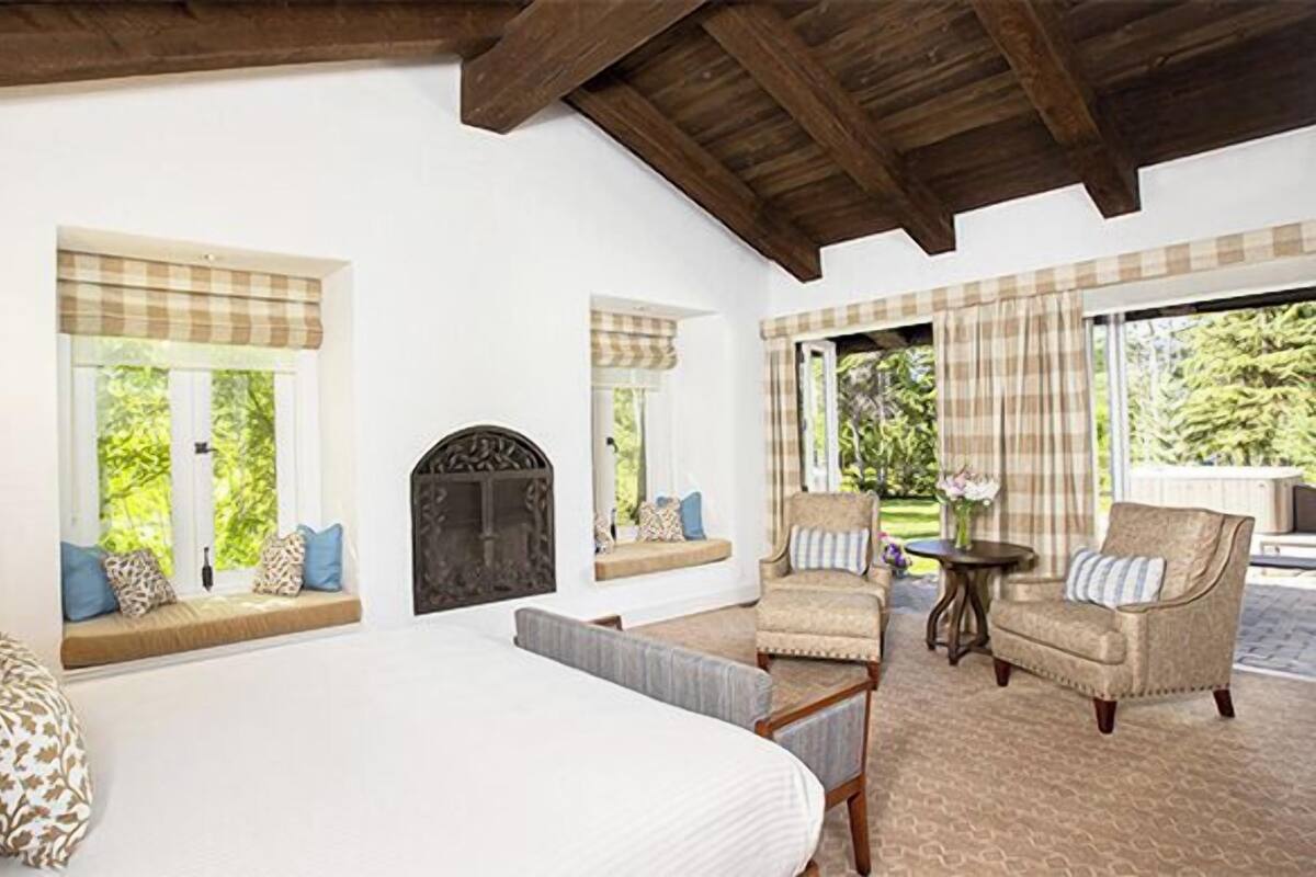 A bedroom with a large bed, two windows with curtains, a fireplace, and a seating area with chairs and a table.