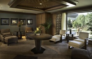A spacious living room with a large round table, several armchairs, and a view of a forest through large windows.