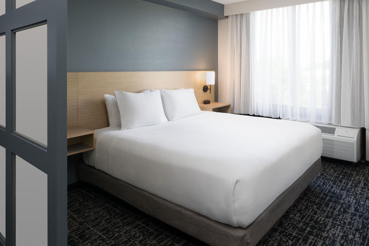 Une chambre d’hôtel avec un grand lit, une table de chevet, une lampe fixée au mur et une fenêtre avec des rideaux légers.