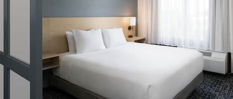 A hotel room with a large bed, a bedside table, a wall-mounted lamp, and a window with sheer curtains.