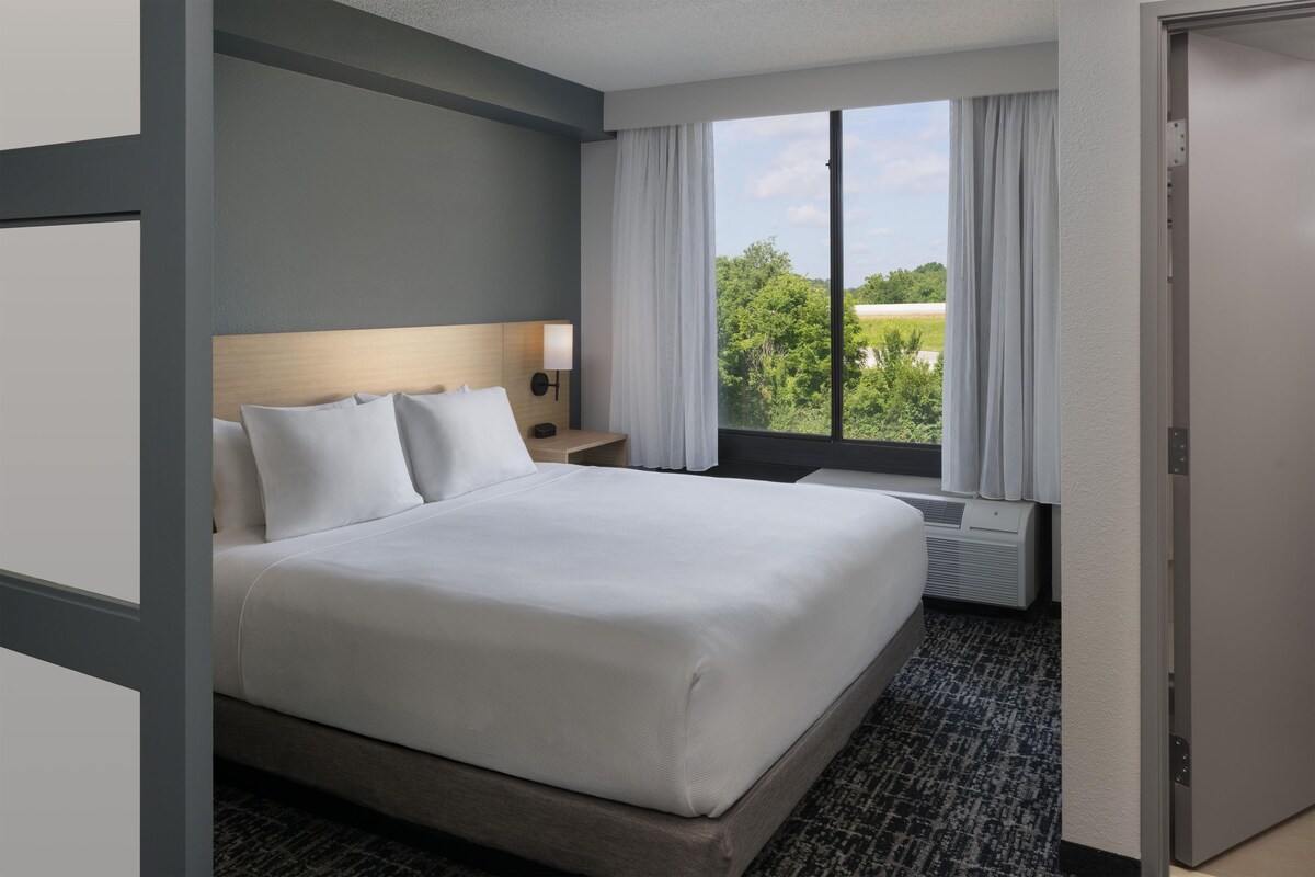 Une chambre d’hôtel avec un grand lit, une table de chevet, une lampe, une fenêtre avec des rideaux et une vue sur la verdure.