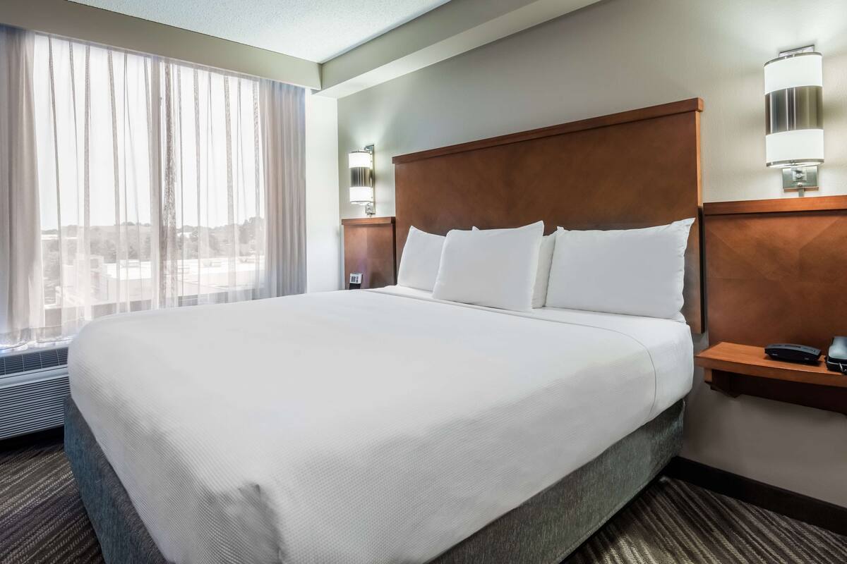 Een hotelkamer met een groot bed, een nachtkastje met een lamp en een raam met gordijnen.