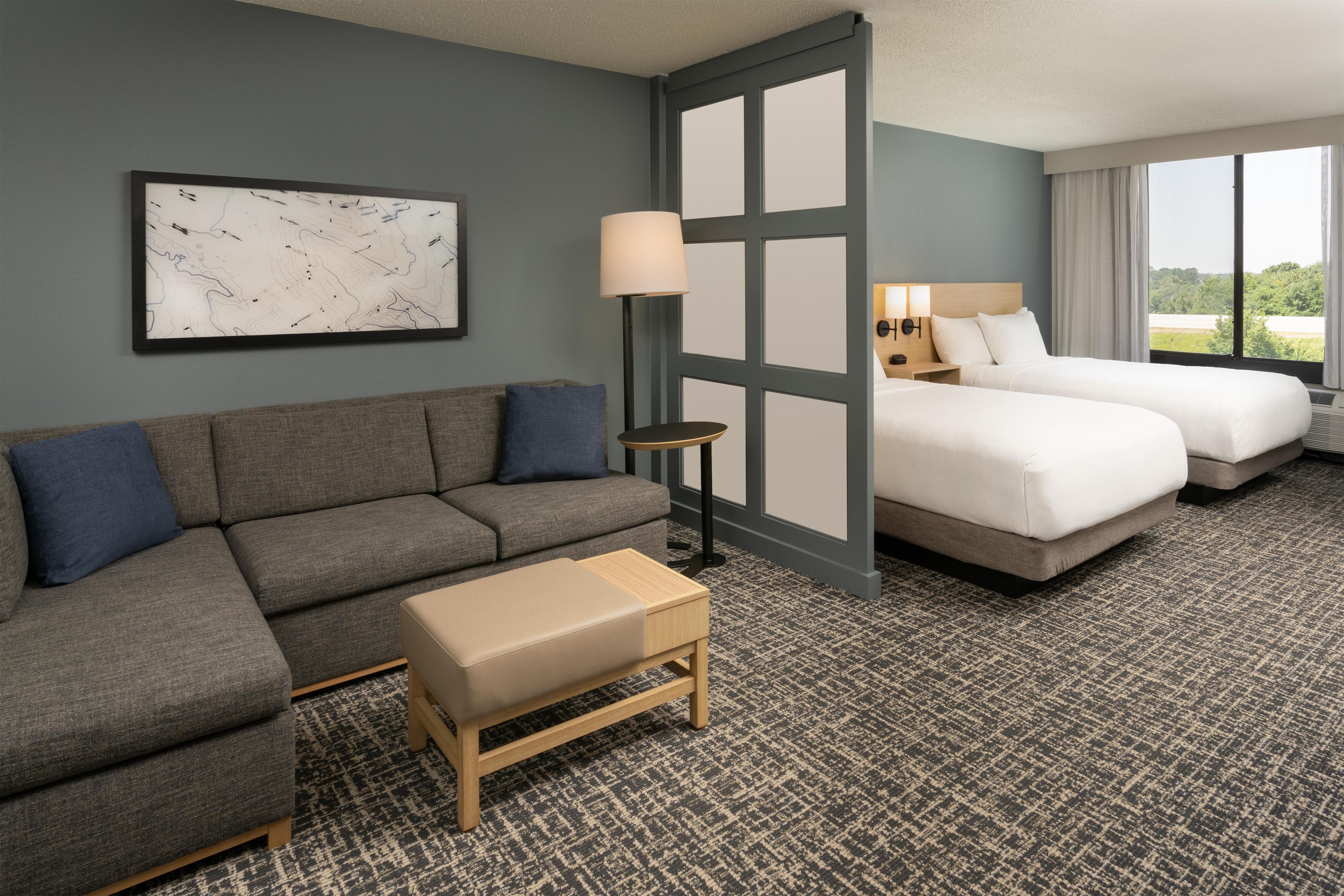 A hotel room with a gray sofa, a small ottoman, two beds, a nightstand, a lamp, and a large window with a view.