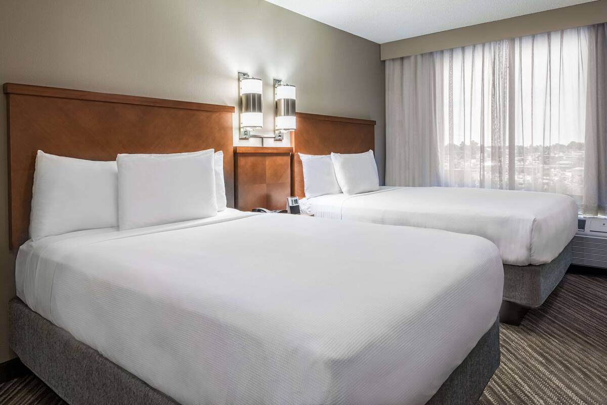 Een hotelkamer met twee bedden, een houten hoofdbord, twee wandlampen en een groot raam met gordijnen.