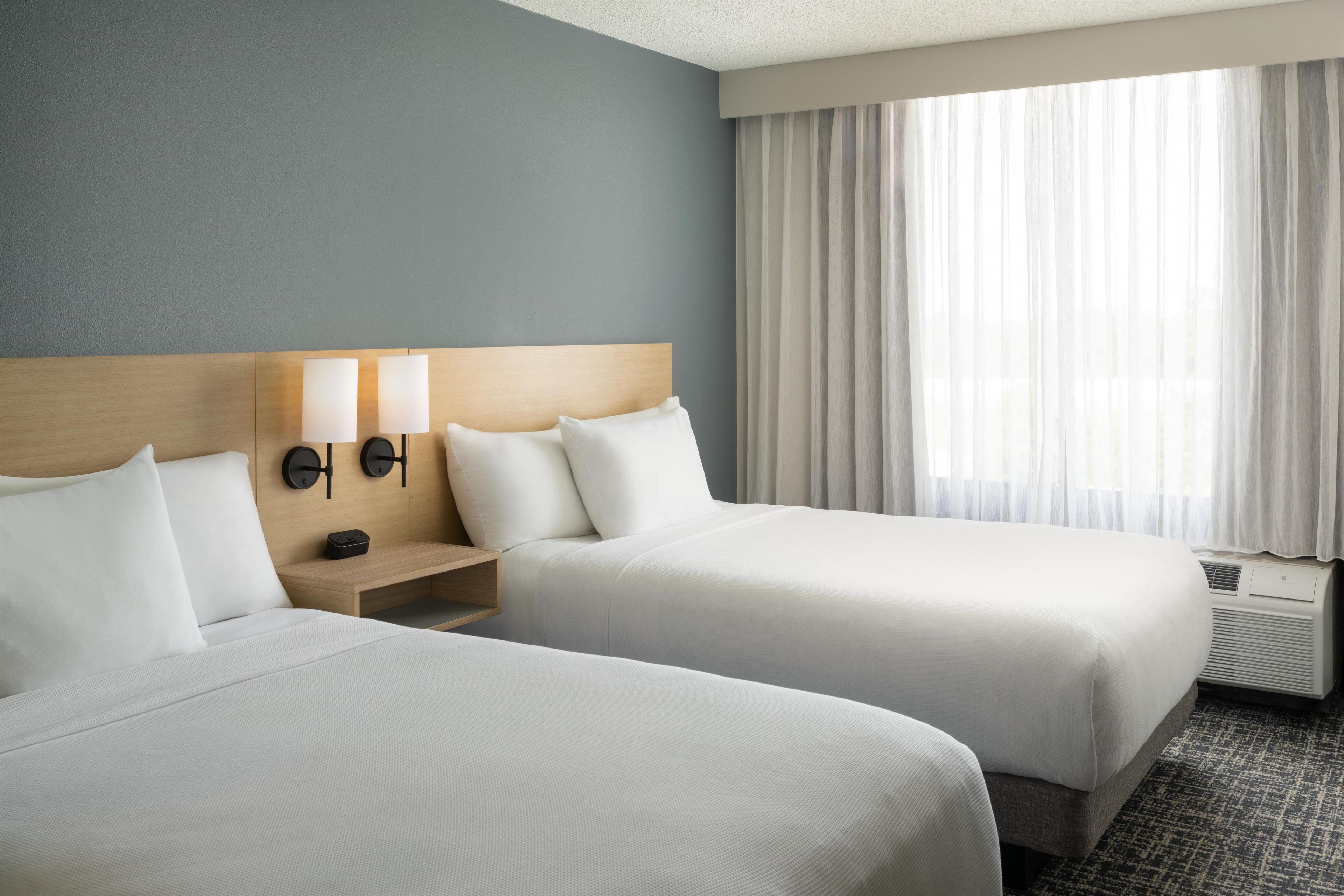 A hotel room with two beds, a nightstand, a wall-mounted lamp, and a window with curtains.