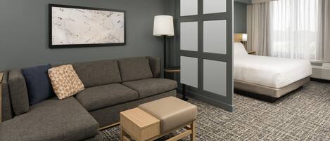 A hotel room with a gray sofa, a small wooden bench, a bed with white bedding, a nightstand with a lamp, and a large window with curtains.